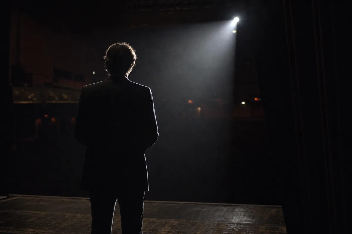 Silhouette d&rsquo;un artiste de dos face à une salle plongée dans la pénombre, faisceau de poursuite latéral