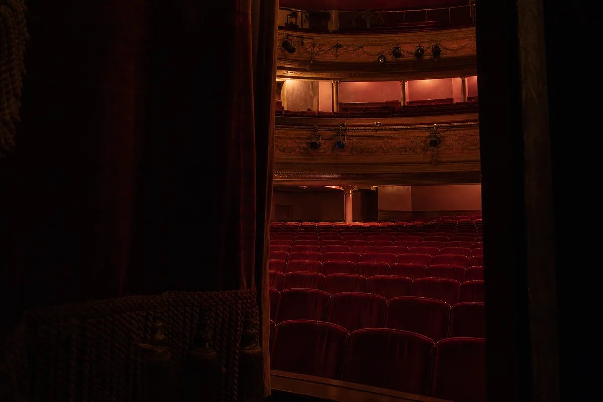 Salle de théâtre vue depuis les coulisses, fauteuils rouges vides et rampe de projecteurs éteinte